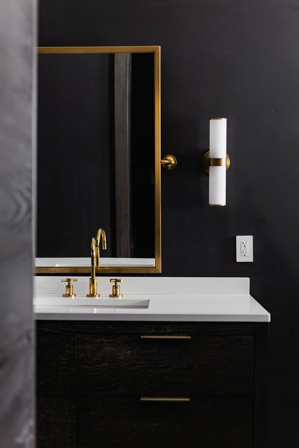 Modern bathroom vanity with gold fixtures, black cabinetry, white countertop, gold-framed mirror, and wall sconce on dark walls.