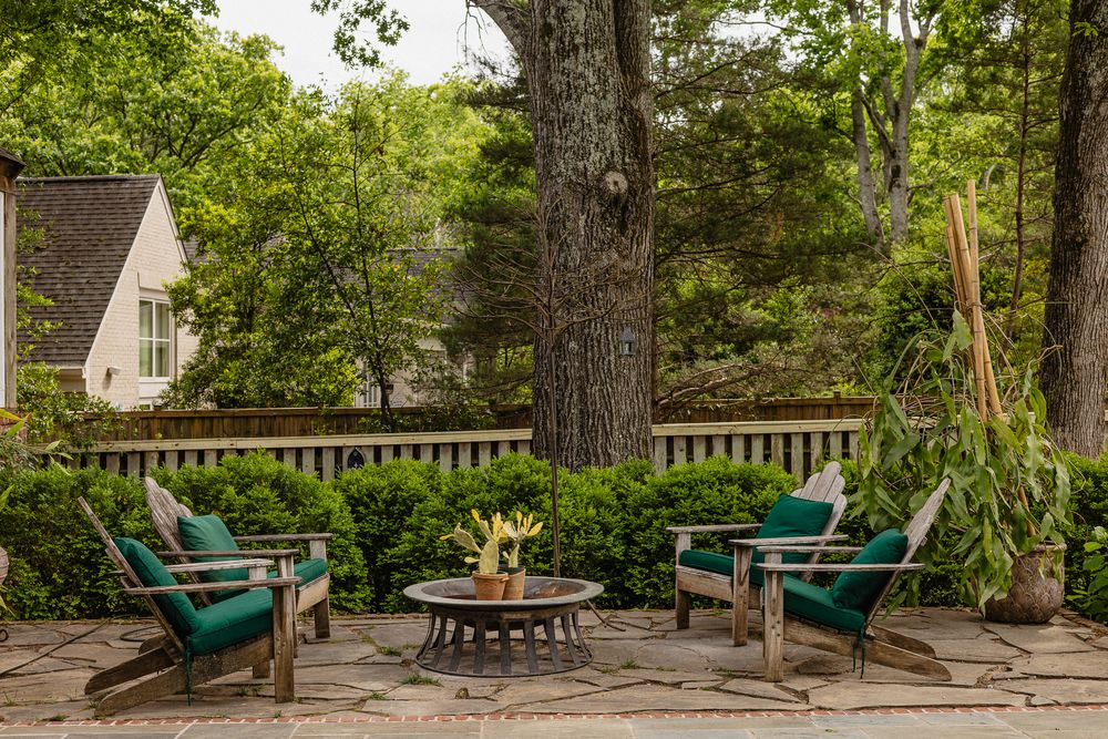 Outdoor patio with Adirondack chairs around a stone fire pit, surrounded by lush greenery and mature trees.
