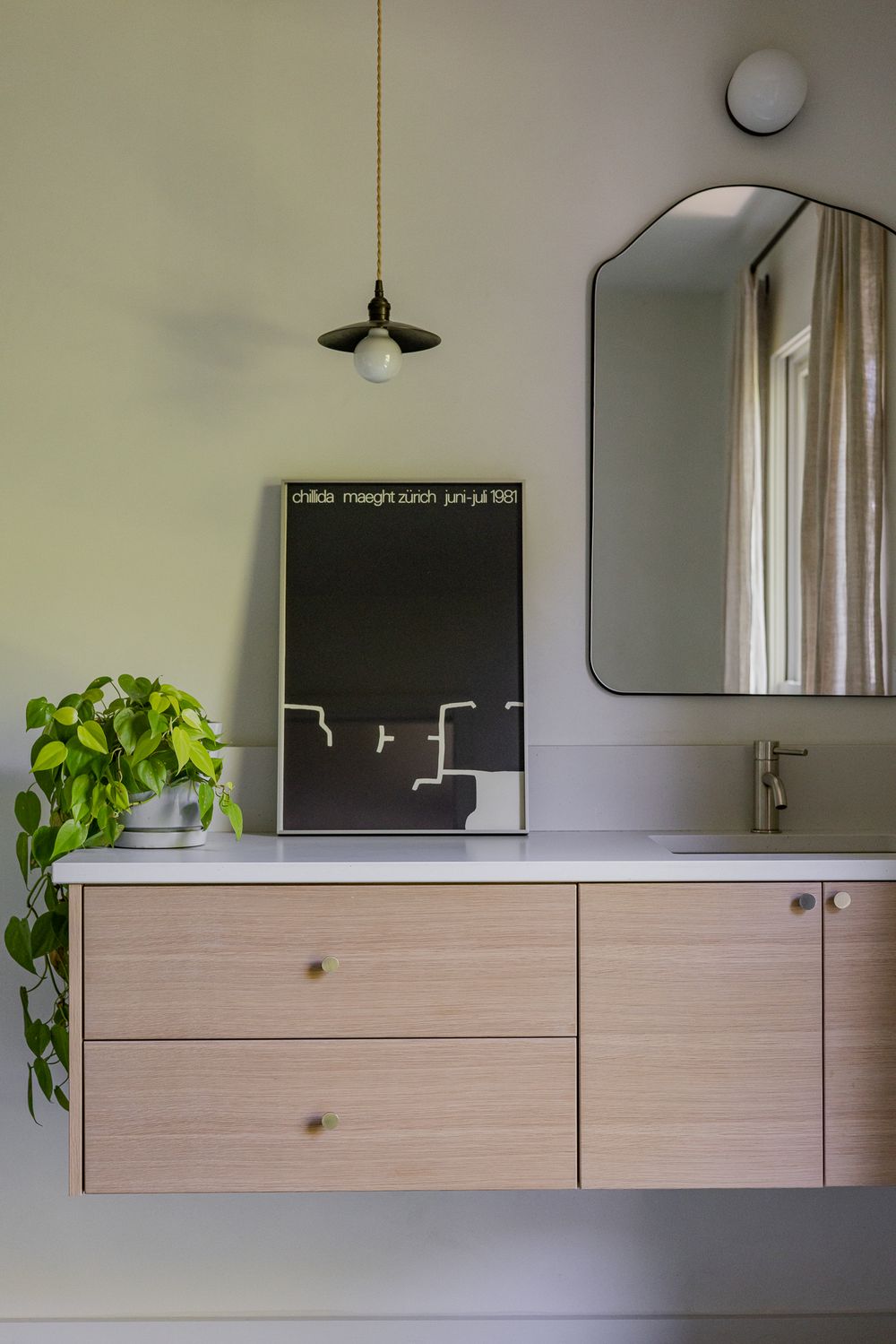 Modern bathroom vanity with wood drawers, black framed mirror, pendant light, green plant, and black art poster on sage green wall.