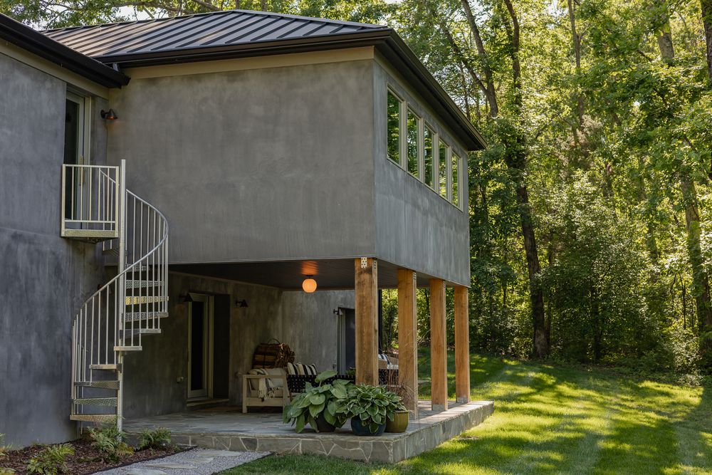 Modern two-story stucco home with spiral staircase, wood pillars, and metal roof surrounded by lush green trees.
