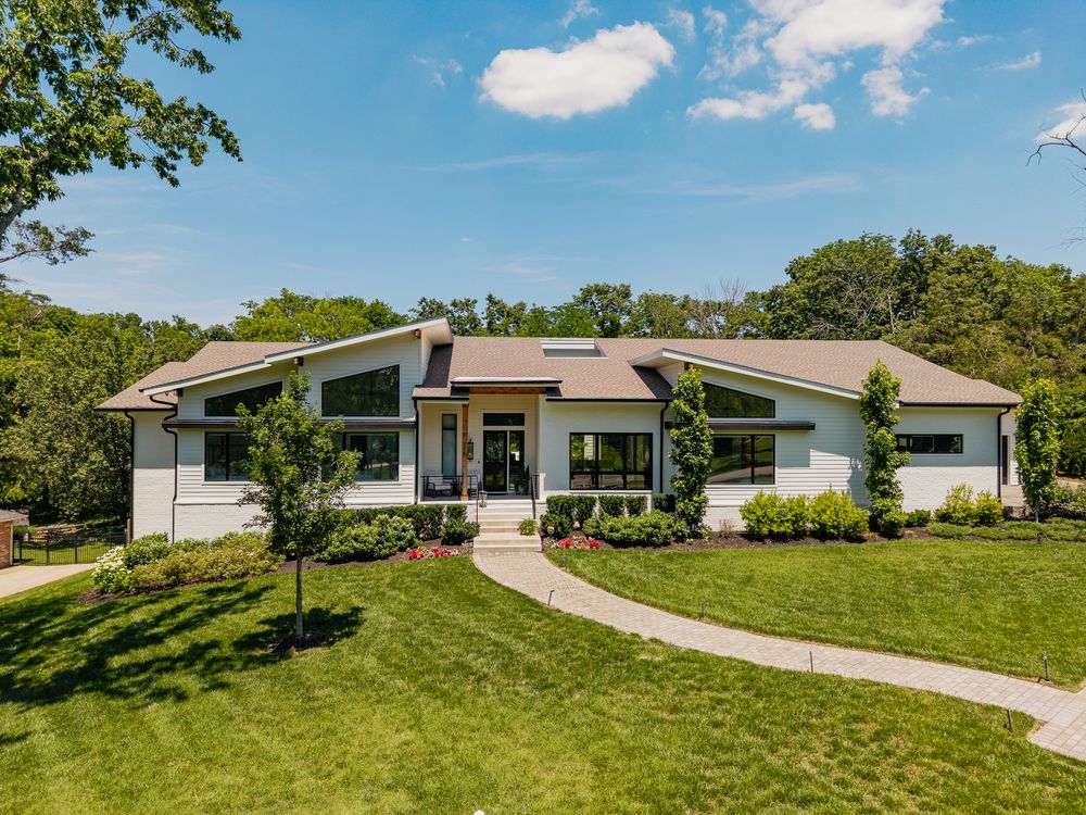 Modern mid-century ranch home with white exterior, large windows, manicured lawn, curved driveway, and lush green trees under blue sky.