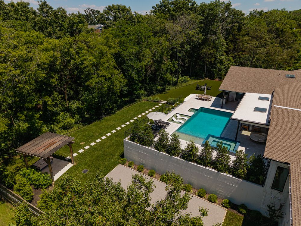 Aerial view of a modern luxury home with a rectangular pool, manicured lawn, and lush tree surroundings.
