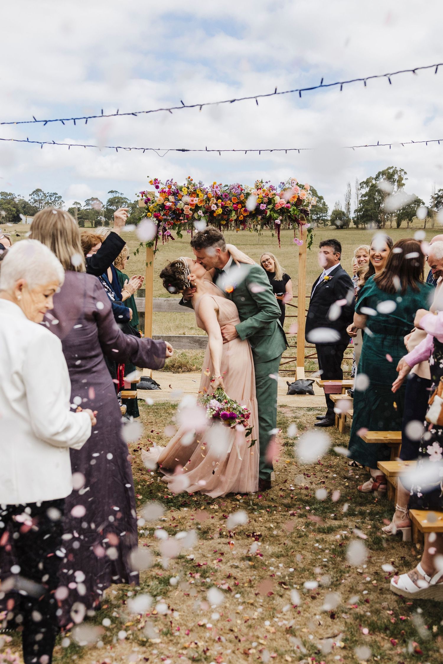 Wedding guests celebrate during an outdoor ceremony with string lights and floral arrangements.