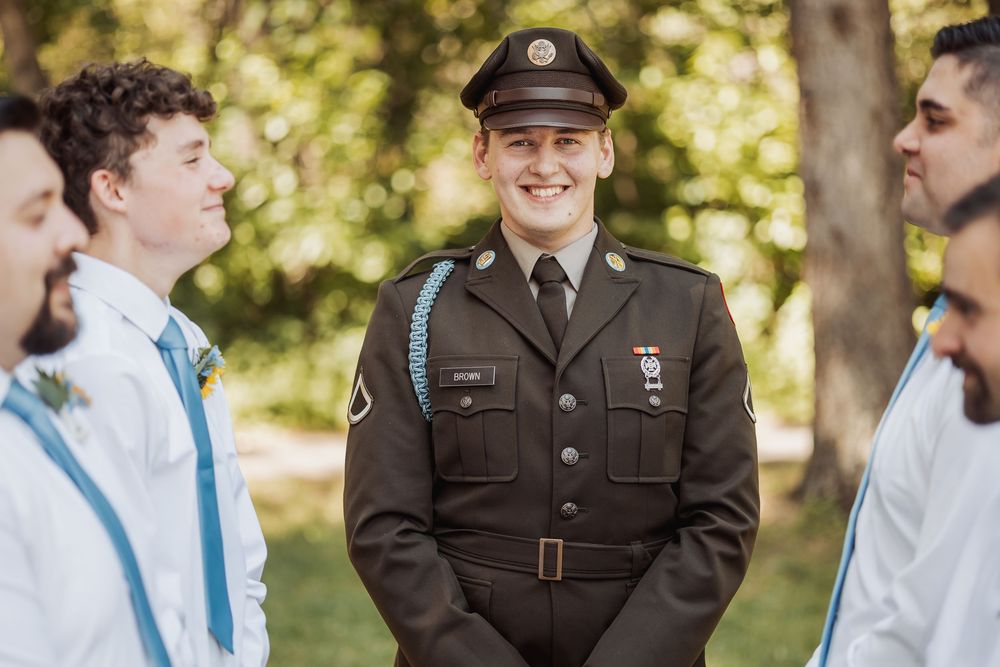 Wedding party poses together outdoors with bridesmaids in white dresses and blue sashes, and groomsmen in dark uniforms.