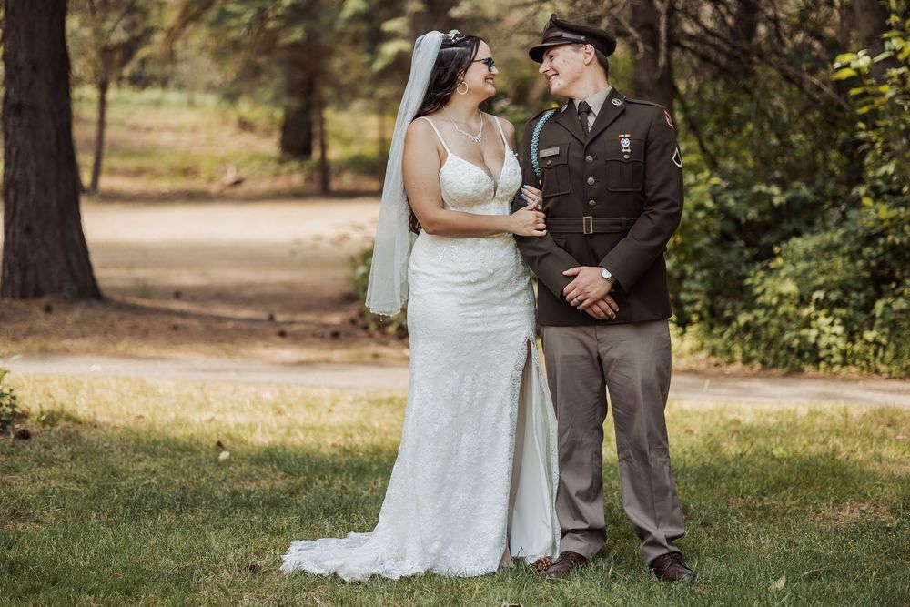 A bride in a white lace gown and military servicemember in dress uniform pose together outdoors in a wooded setting.