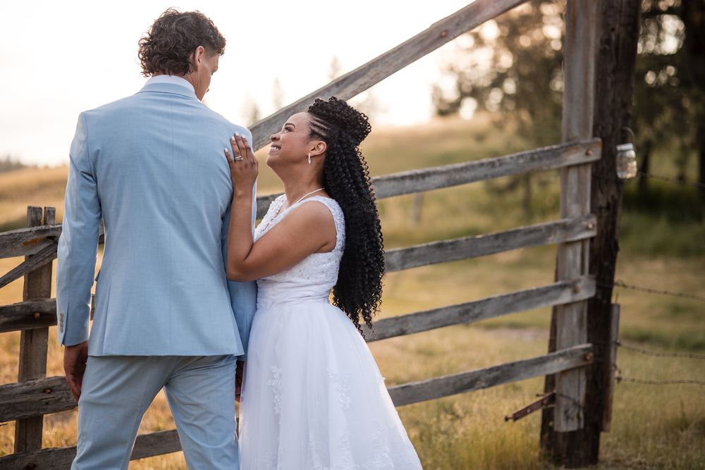 A couple shares a romantic moment together at sunset near a rustic wooden fence in a rural setting.