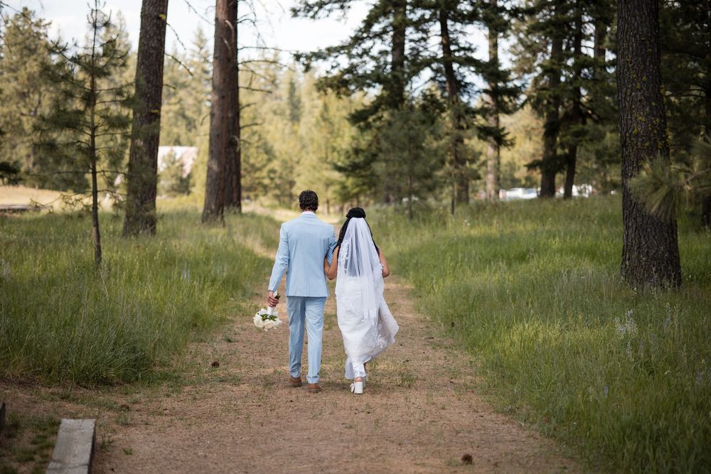 Two people in light-colored outfits walking together along a dirt path through a wooded area on a sunny day.