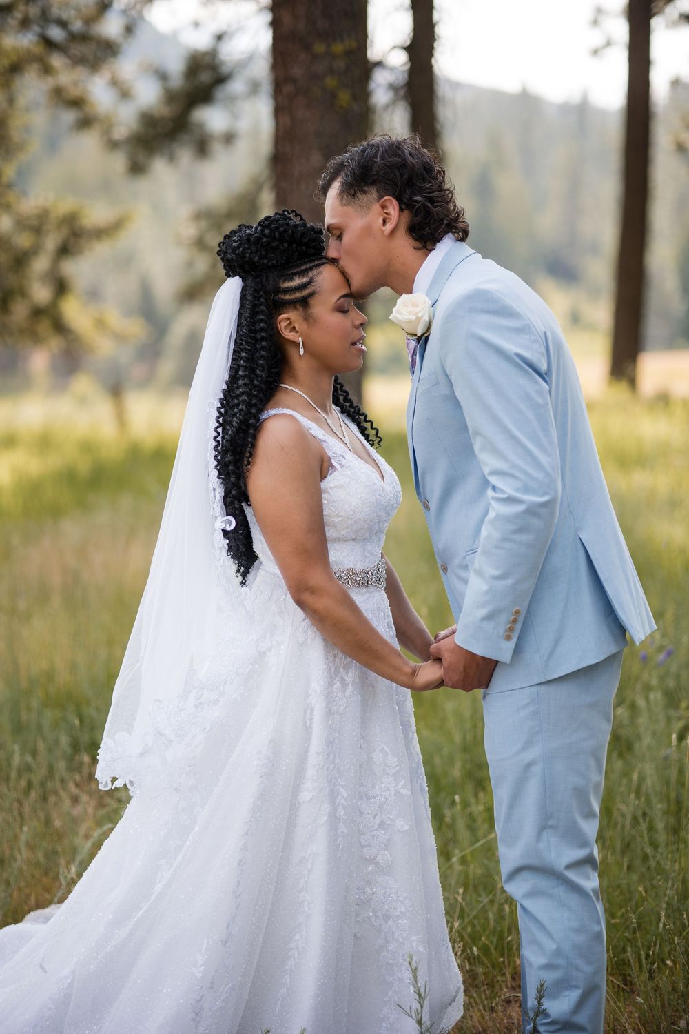 Newlywed couple shares an intimate moment outdoors in a grassy field with the bride in white and groom in light blue suit.
