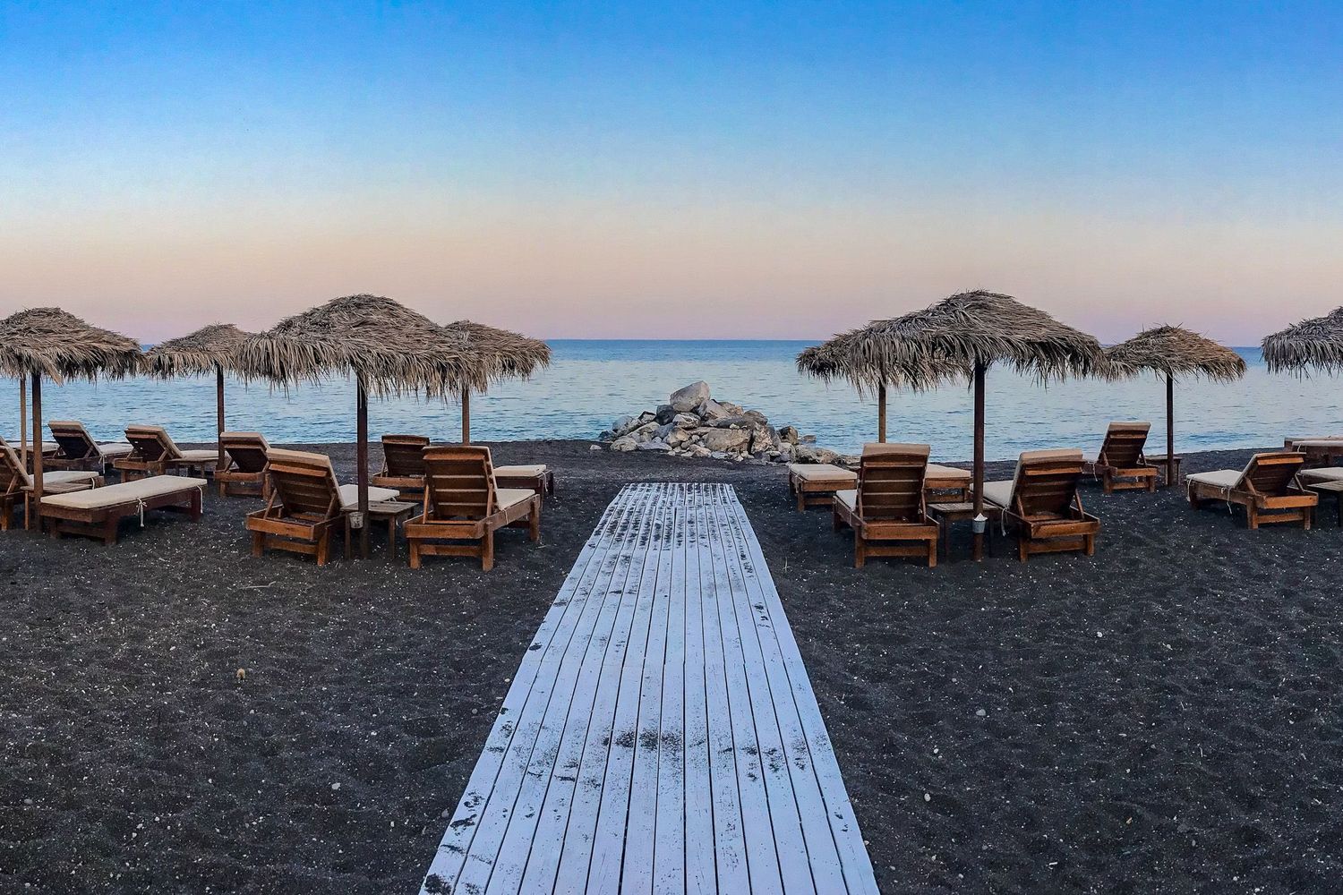 Santorini Greece travel photography of straw umbrellas and wooden lounge chairs on a black sand beach with a wooden path to the ocean.