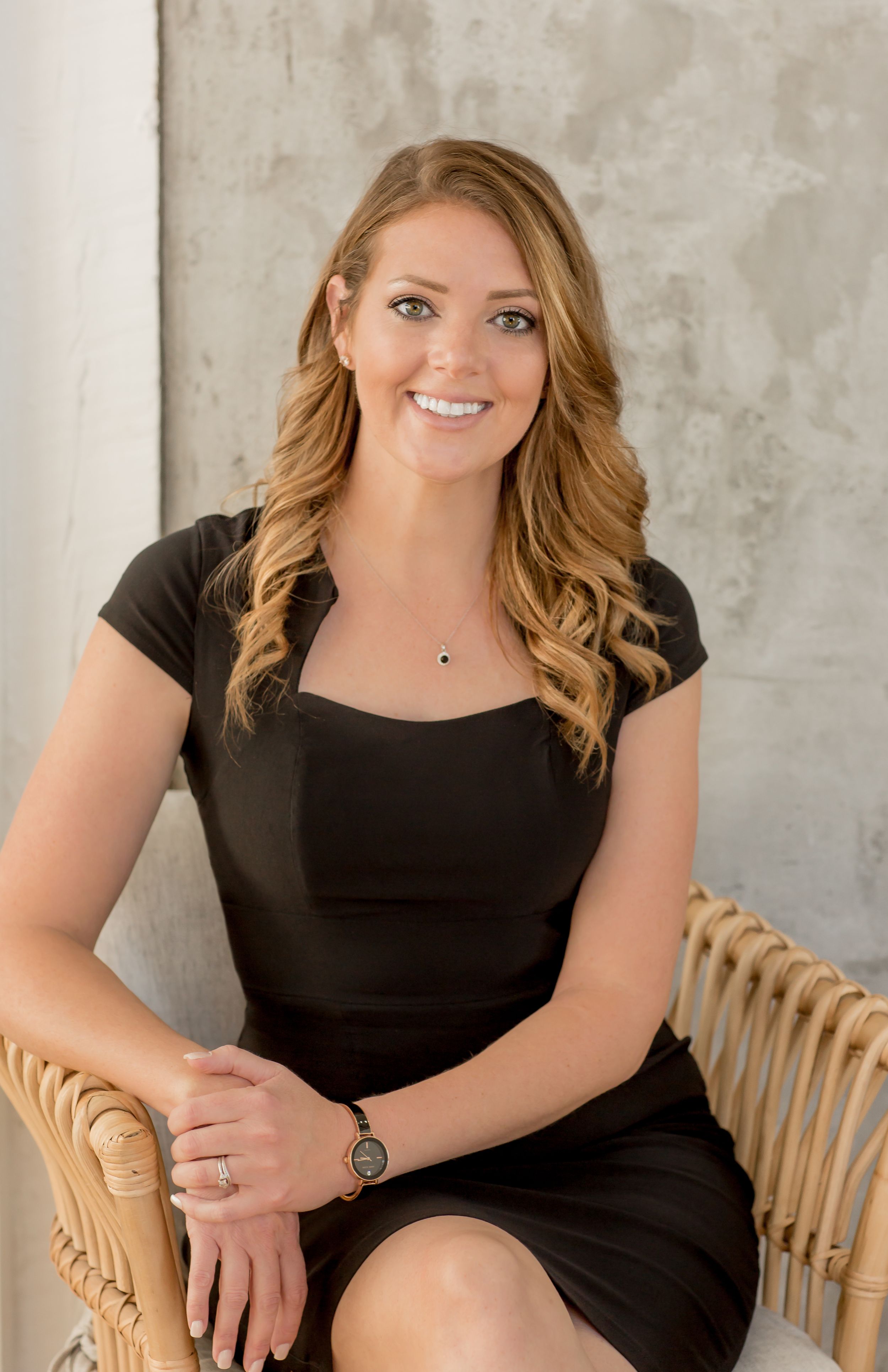 Professional woman in black dress sitting in wicker chair, smiling confidently against neutral backdrop.