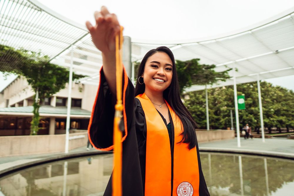 Graduate in orange graduation gown and black robe raises diploma while standing near fountain at outdoor ceremony.