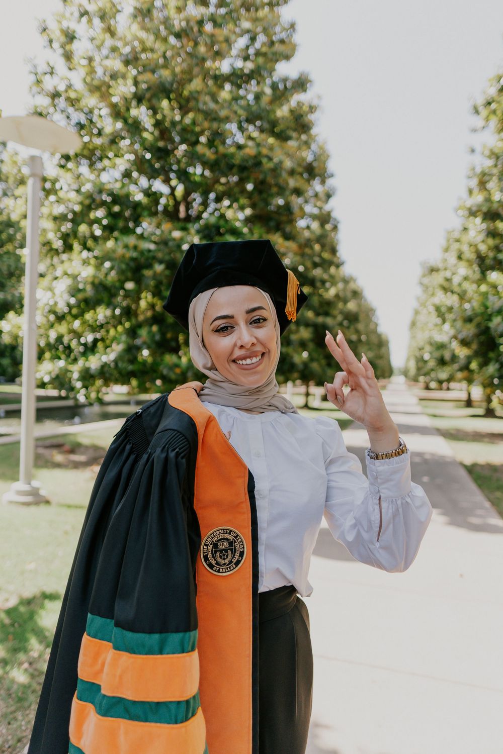 Graduate in academic regalia with orange and green stole poses outdoors on sunny day during commencement.