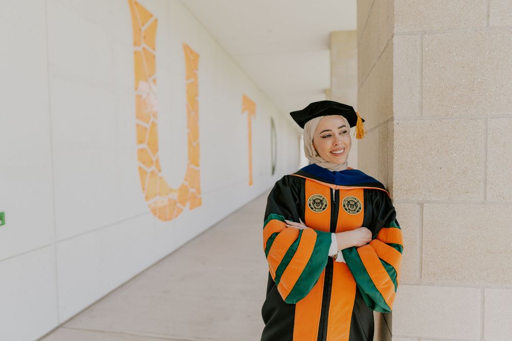 Graduate in orange and black academic regalia stands in a hallway during commencement ceremony.