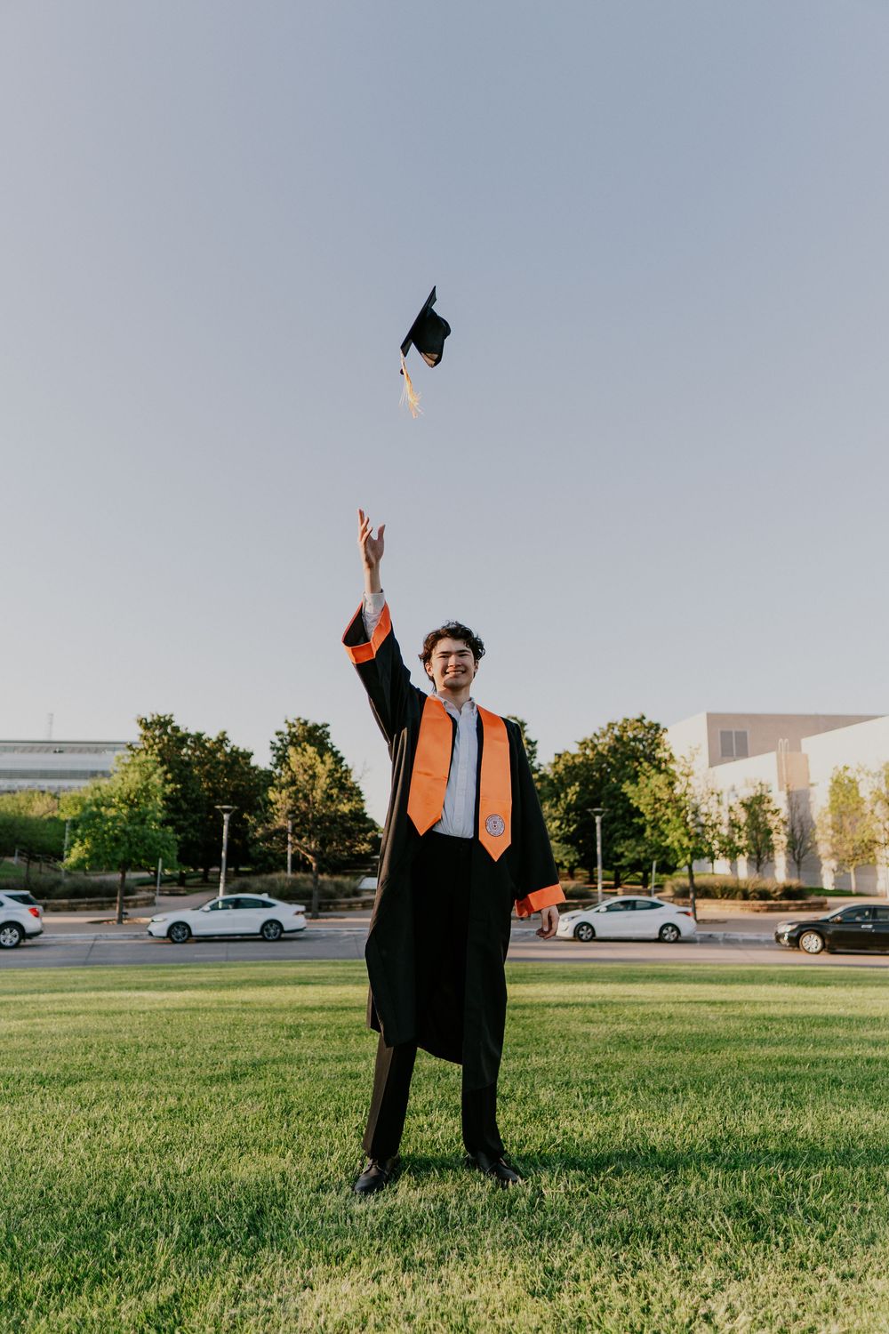 Graduate in cap and gown tosses graduation cap in the air while standing on grass with cars in background.