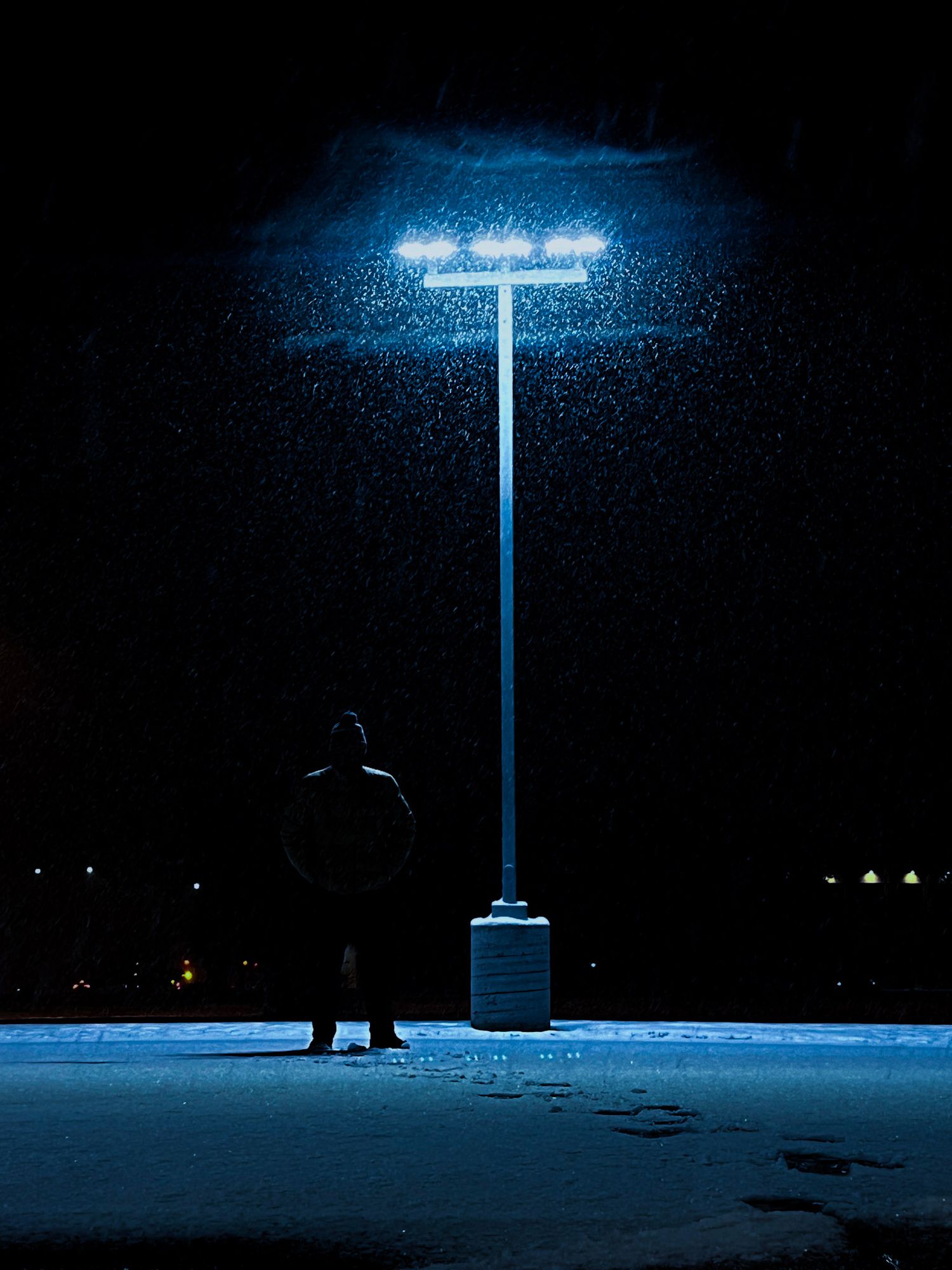 A solitary street lamp casts an eerie blue glow on snowy ground at night, creating a moody winter atmosphere.
