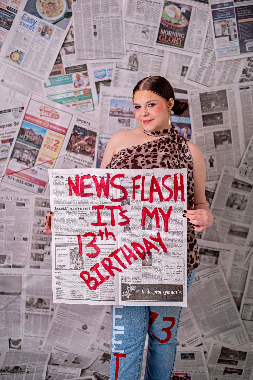 Teen girl holds newspaper sign reading News Flash Its My 13th Birthday, standing before a wall of newspapers.