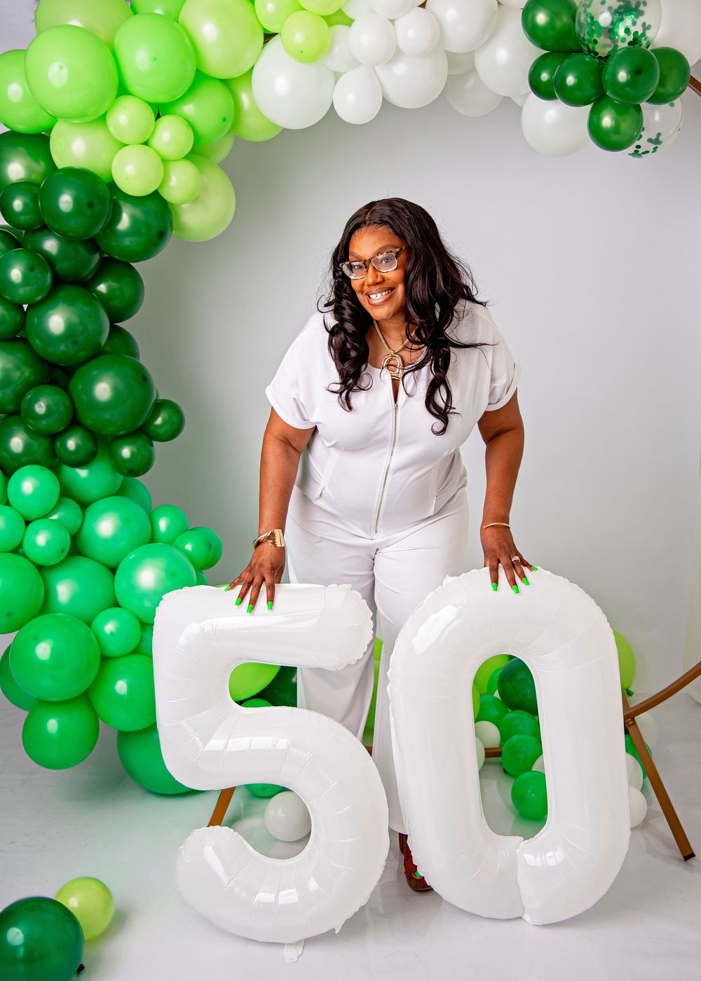 Woman celebrating her 50th birthday posing with large white number balloons and green balloon garland backdrop.