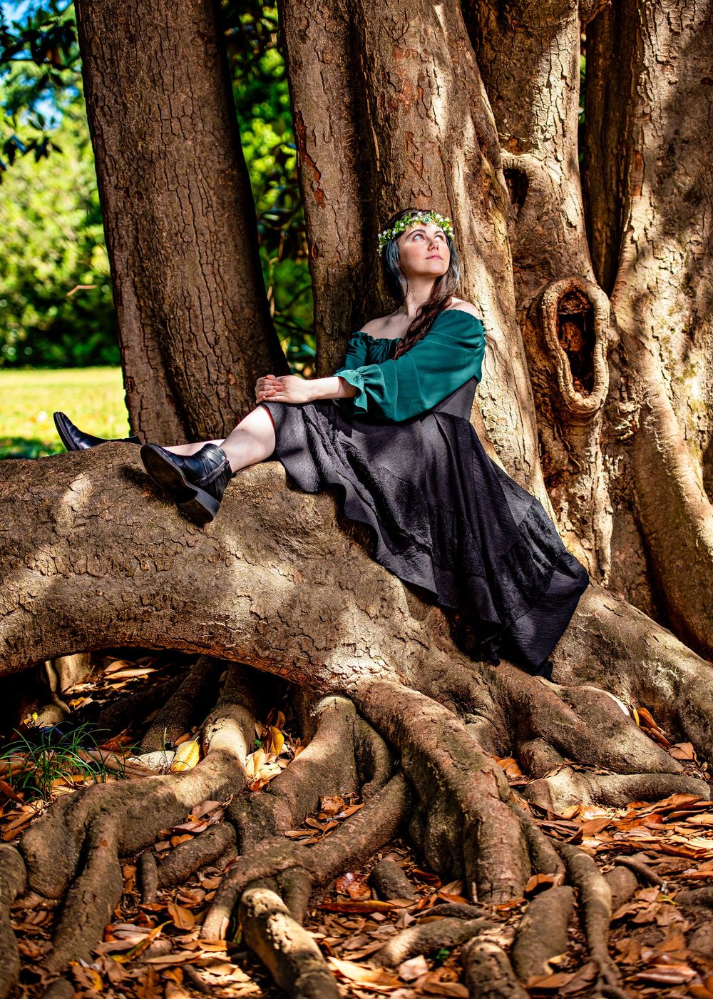 Woman in green blouse and black skirt with floral crown sits on large tree roots in a lush park setting.