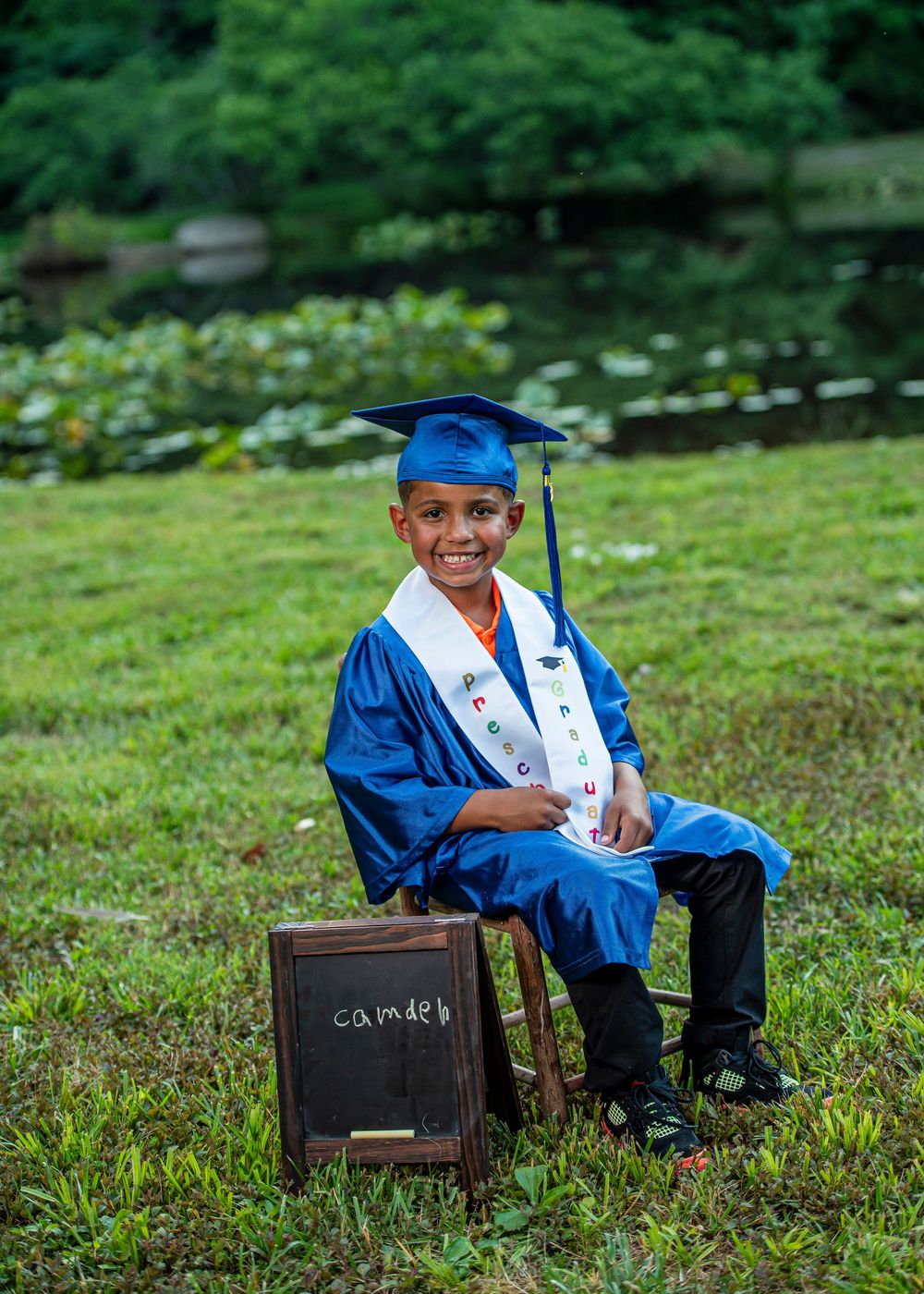 Young boy in blue graduation cap and gown sits on chair outdoors with chalkboard name sign, smiling proudly.
