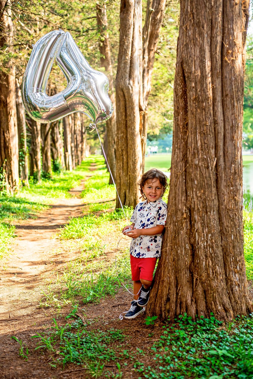 A young boy leans against a tree in a forest path, holding a silver number 4 balloon, wearing red shorts and a white shirt.