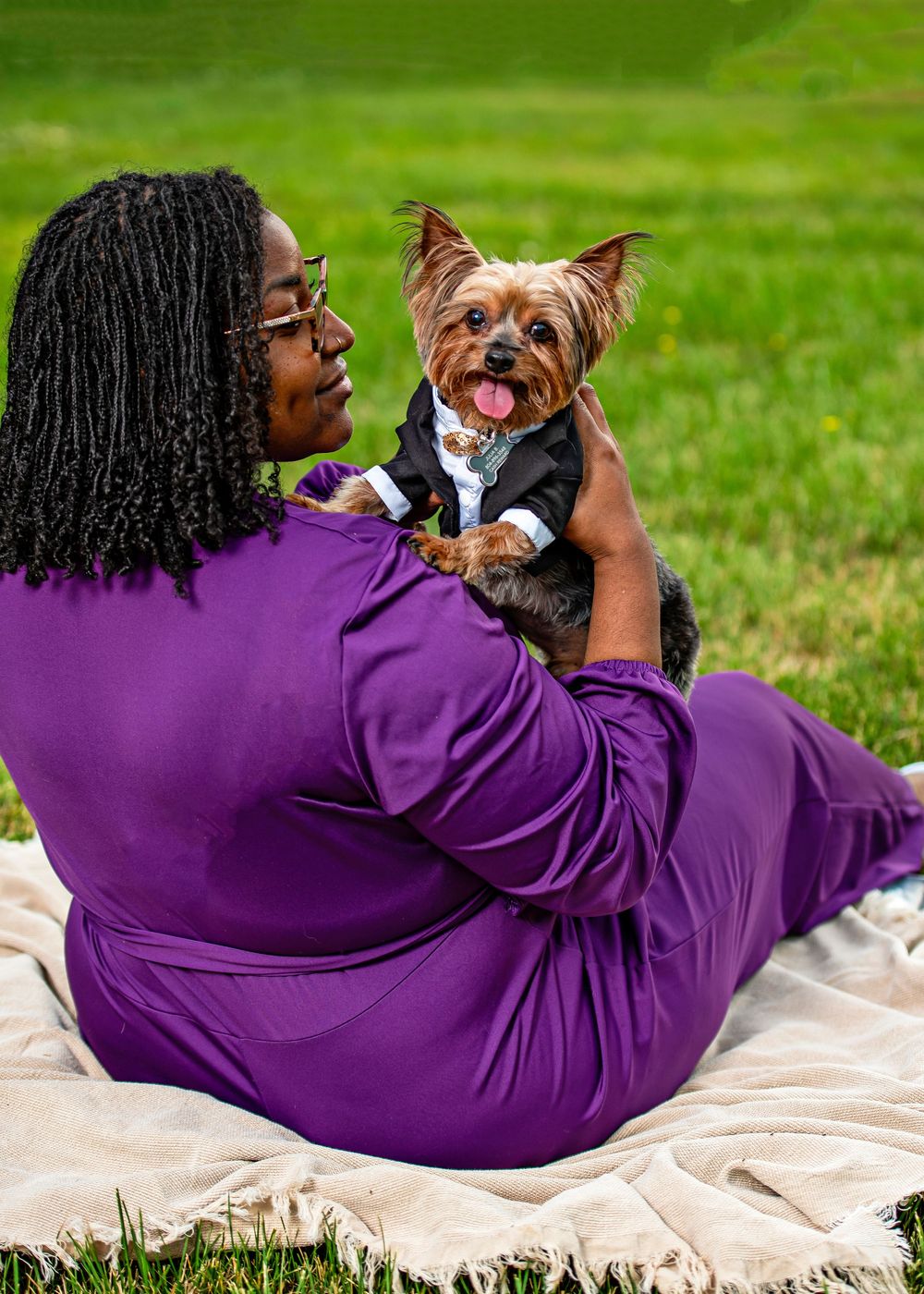 Woman in purple dress sits on blanket holding a Yorkshire Terrier in a tuxedo outfit on a green lawn.