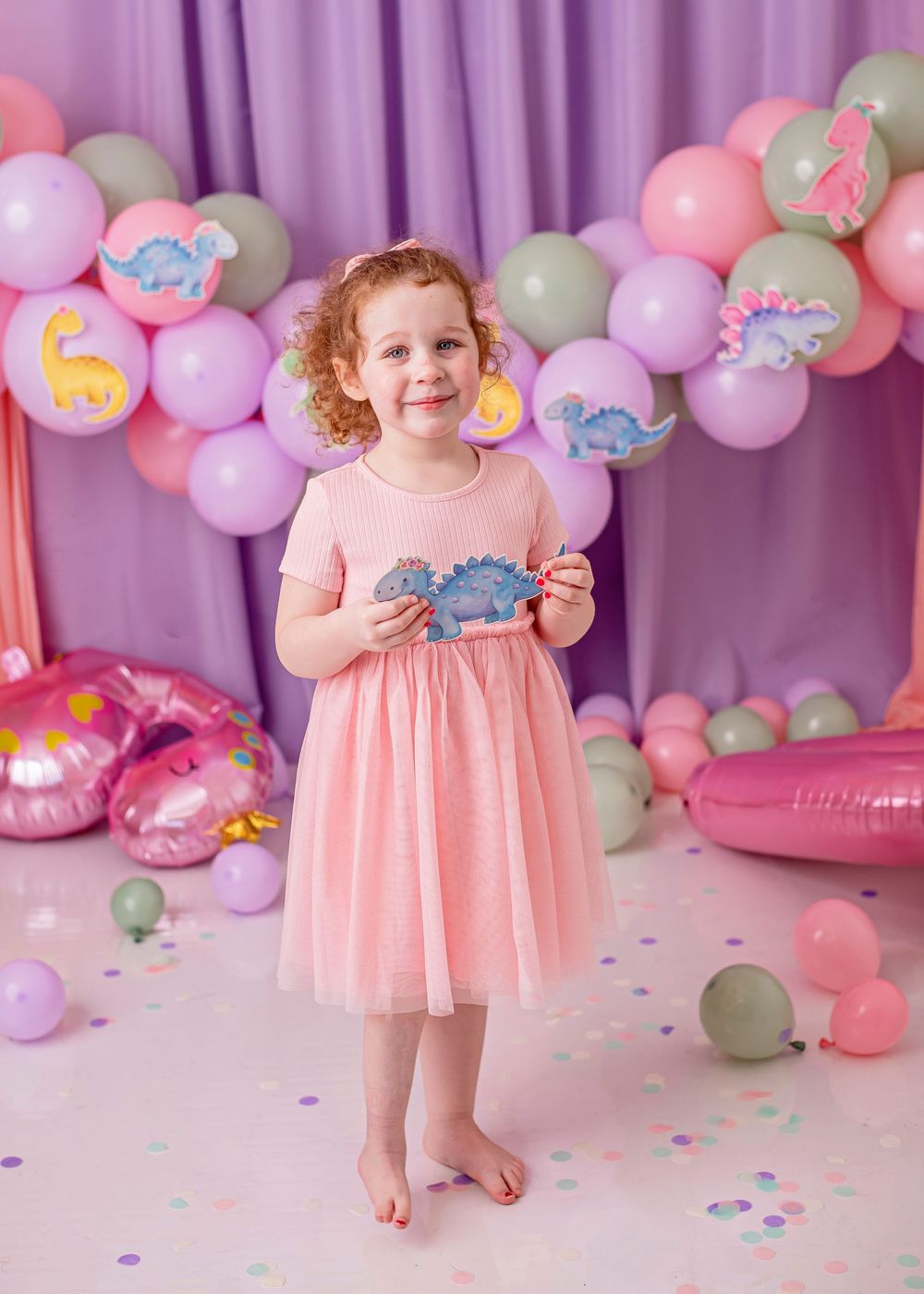 A toddler in a pink dress plays among pastel balloon decorations and confetti at a festive party setup.