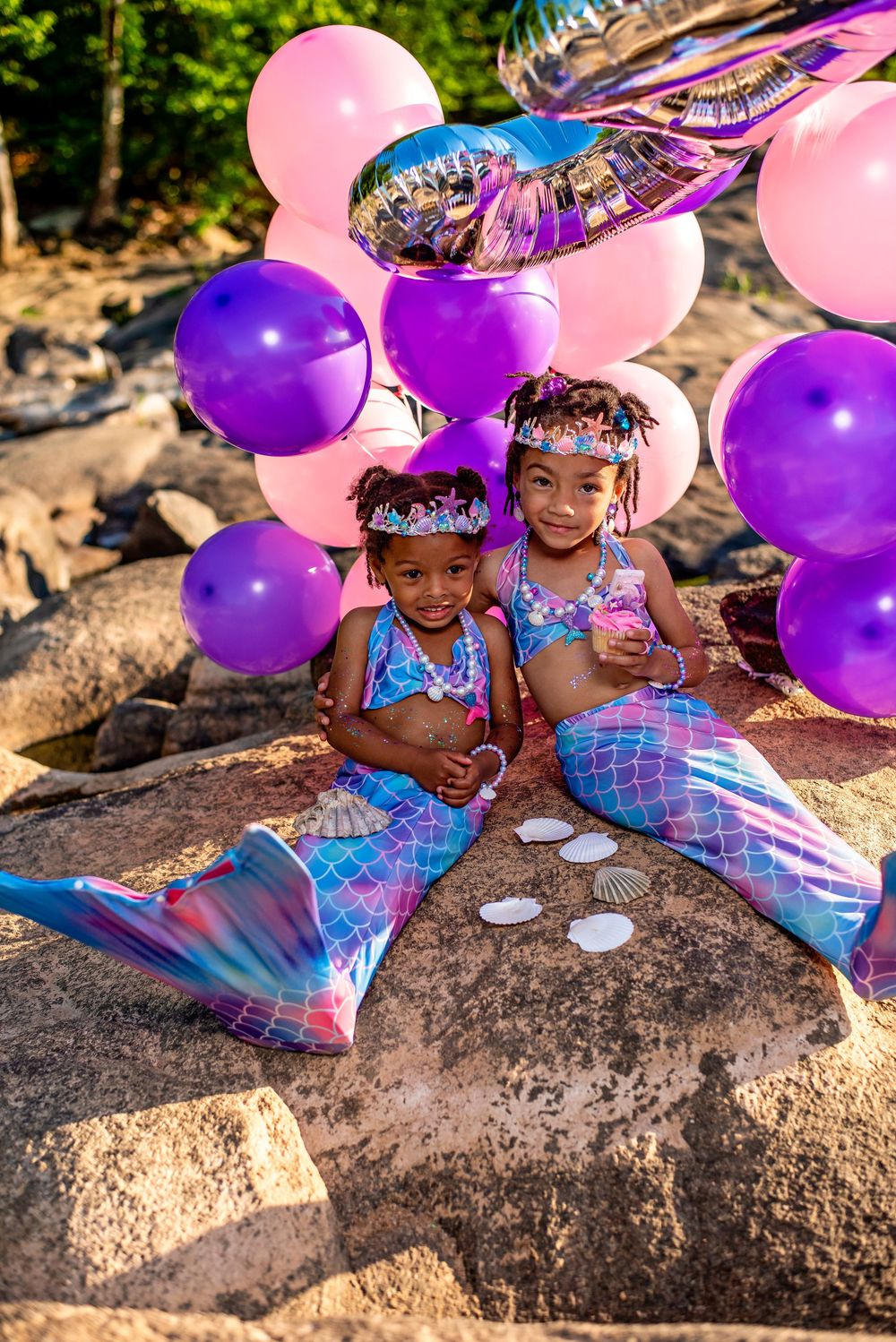 Two young girls in mermaid costumes with tails and tiaras pose on rocks surrounded by pink and purple balloons.