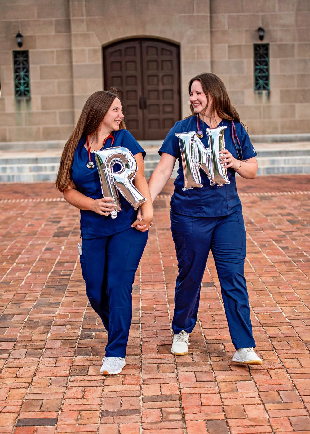 Two nurses in blue scrubs celebrate RN graduation holding silver letter balloons spelling RN on a brick plaza.