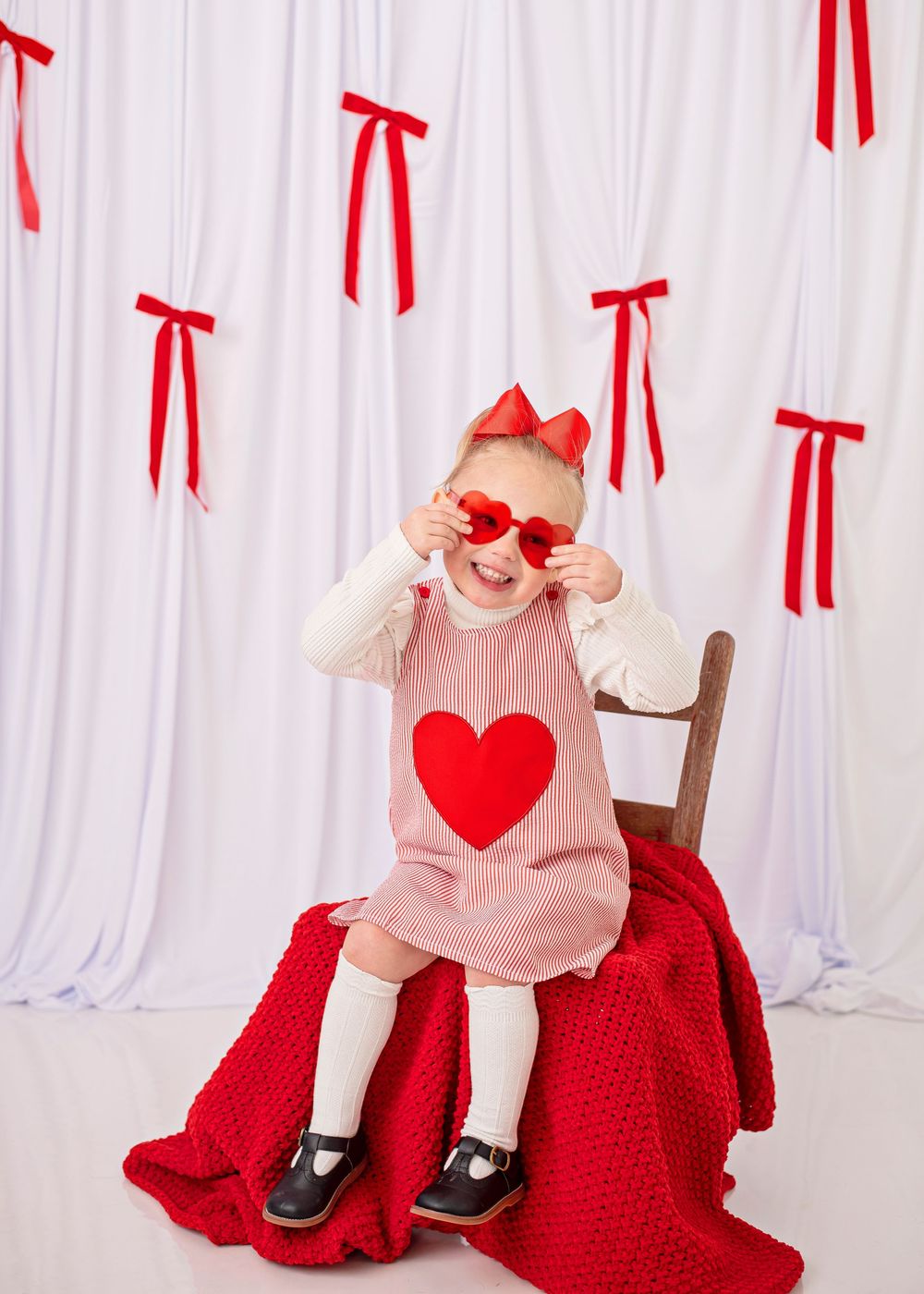 Baby wearing pink dress and red heart sits on red blanket with Valentine's Day themed decorations and ribbons.