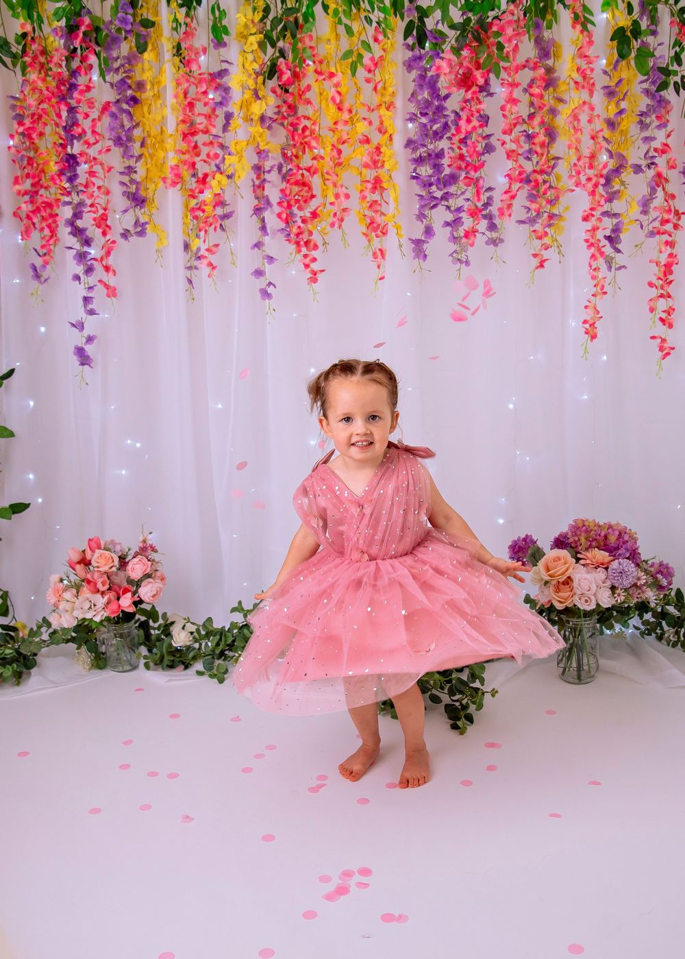 A child in a pink tulle dress poses in front of a floral backdrop with hanging wisteria flowers and twinkling lights.