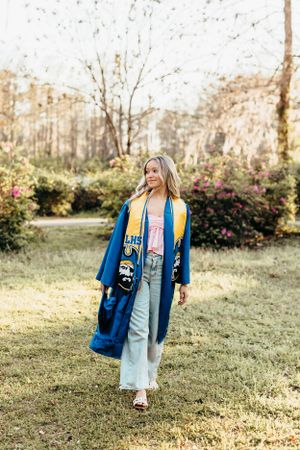 Woman in a blue graduation gown poses in a garden setting with blooming flowers and trees.