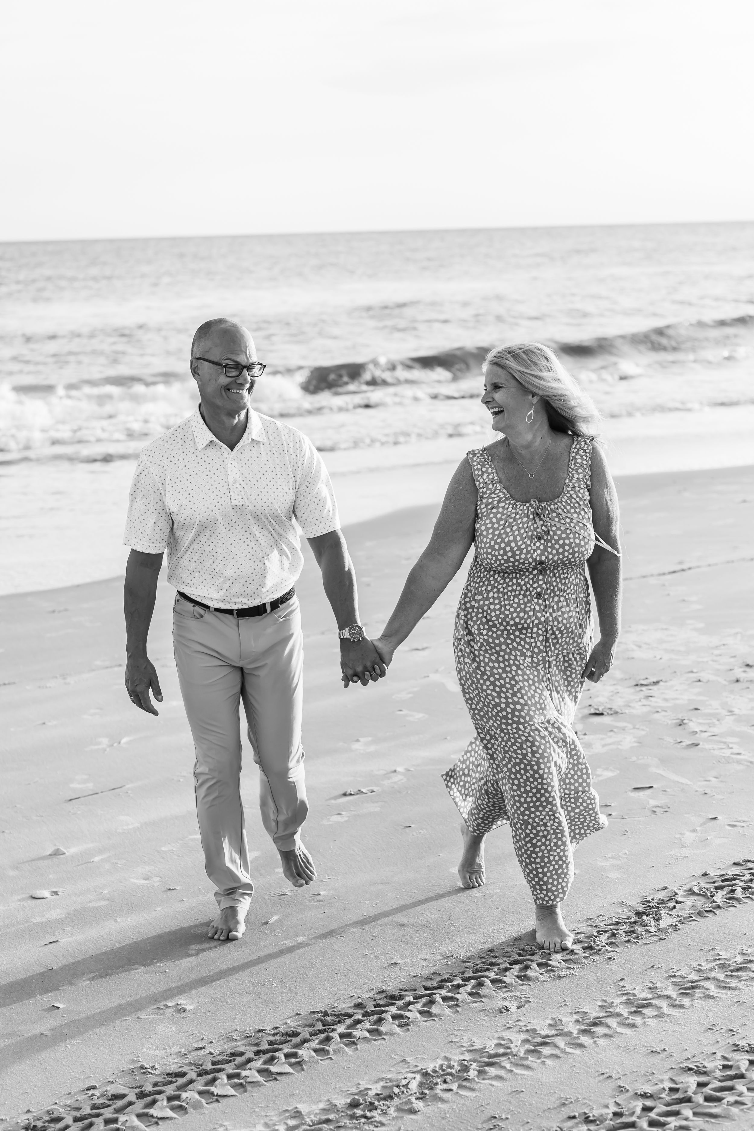 Black and white photo of a happy couple holding hands and walking barefoot on a sandy beach with ocean waves behind them.