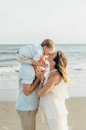 A family shares a tender moment on the beach, with parents kissing their toddler son by the ocean.