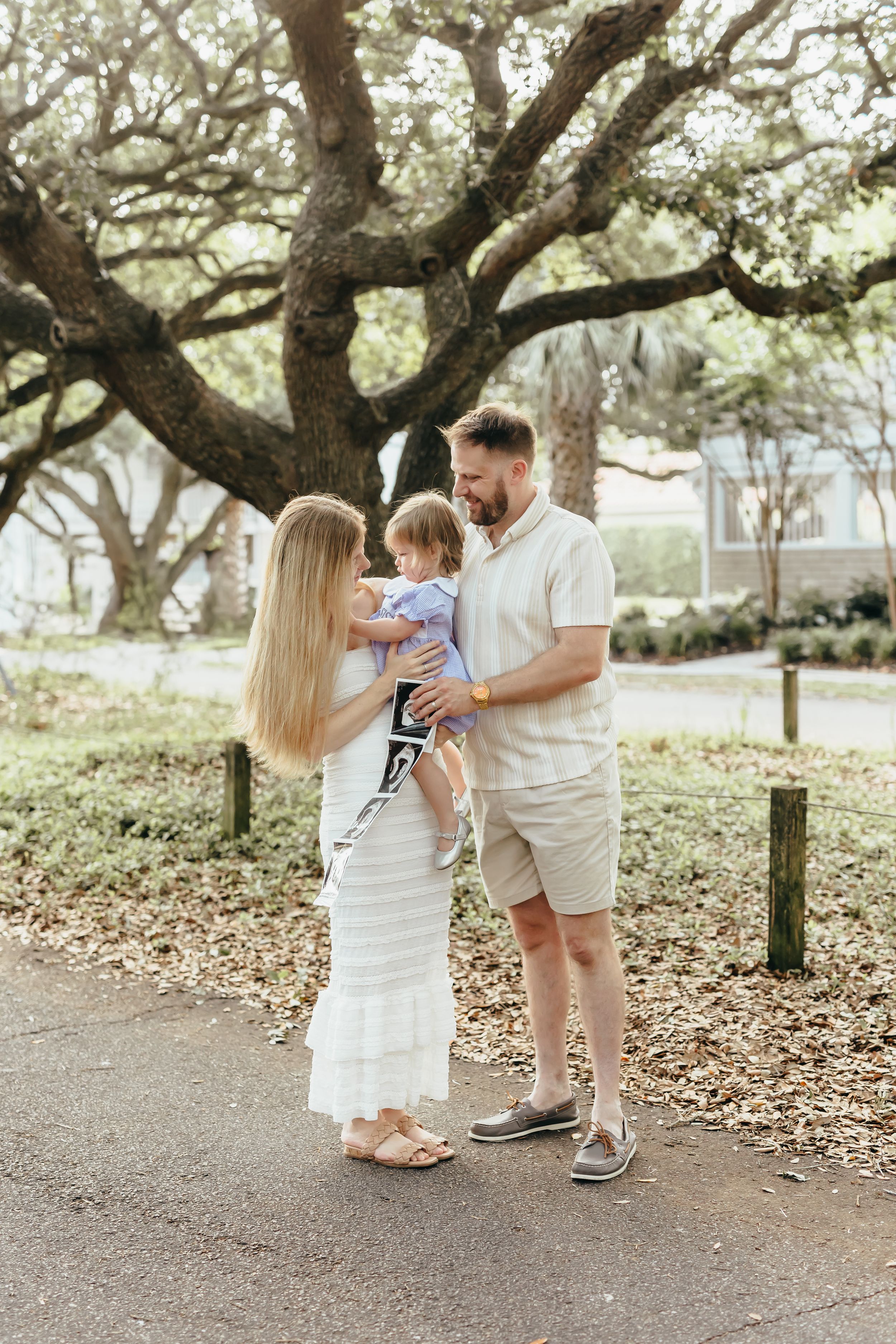 A young family stands outdoors under oak trees, parents holding toddler and ultrasound photo, announcing pregnancy.