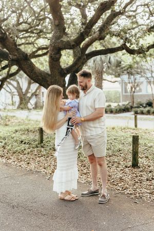 A young family stands outdoors under oak trees, parents holding toddler and ultrasound photo, announcing pregnancy.