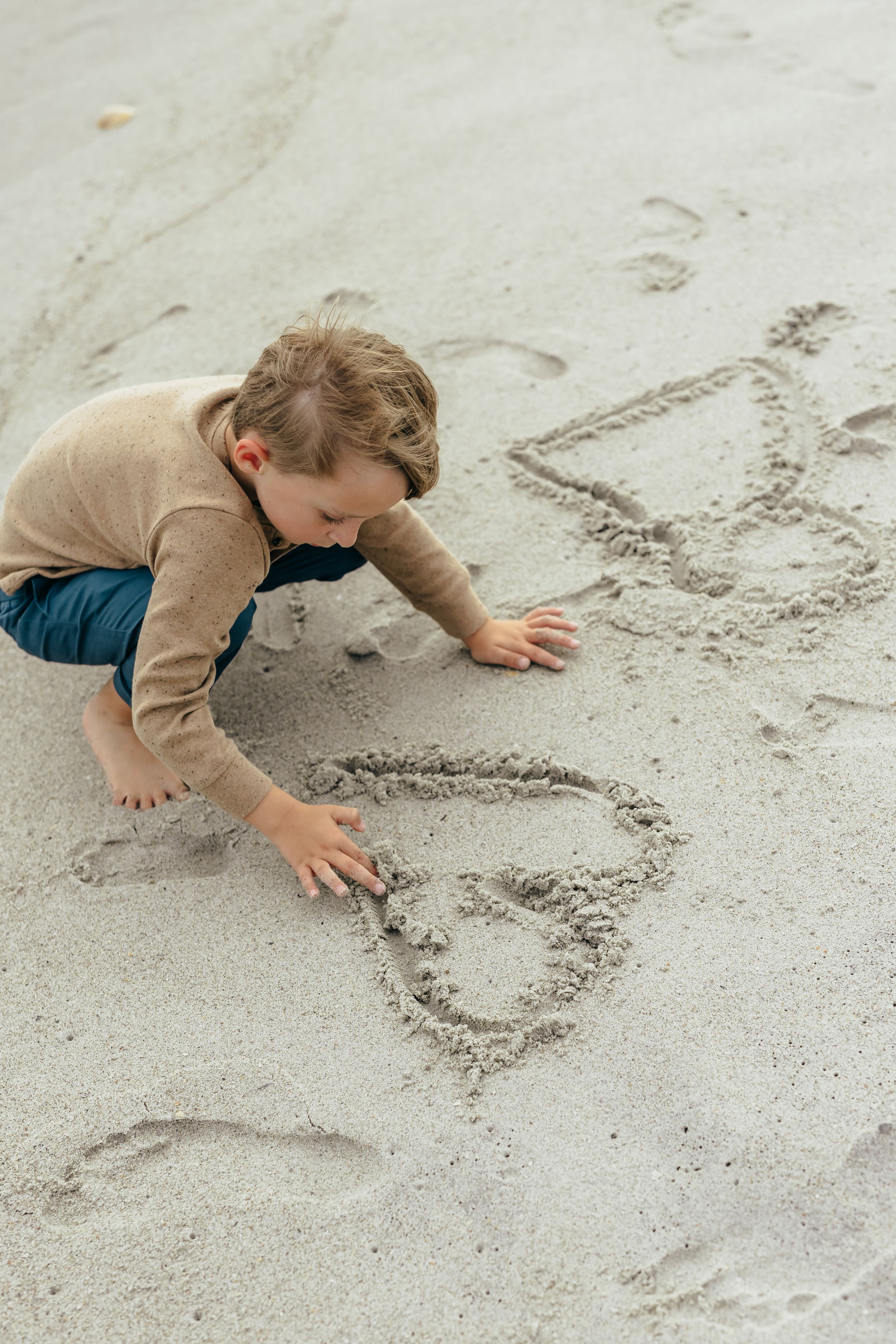 Young child drawing hearts in the sand at the beach, wearing a beige sweater and blue pants.