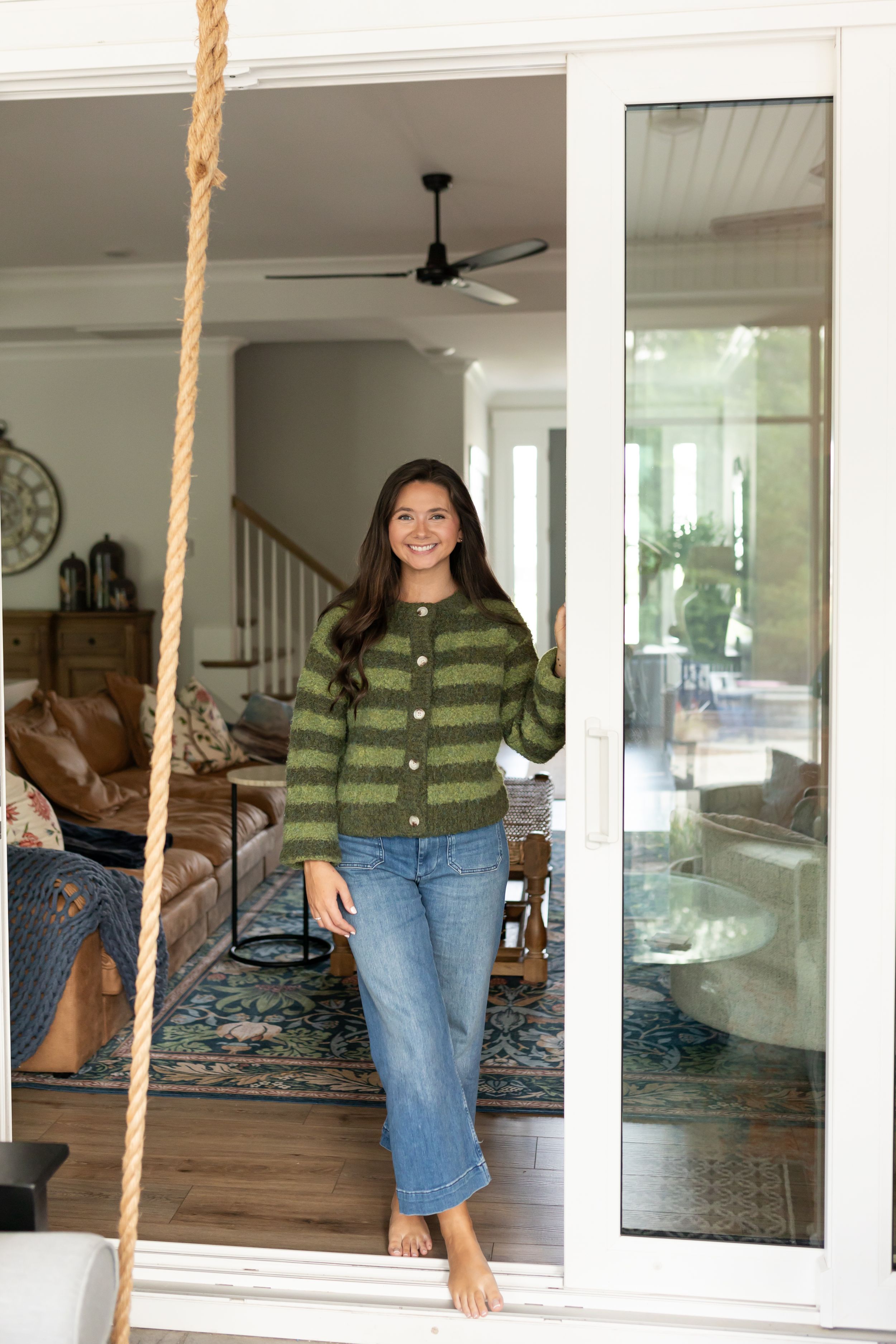 Woman in green striped cardigan and jeans standing in sliding glass doorway of cozy home interior.
