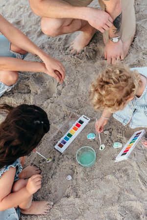 Family painting rocks with watercolors on sandy beach, children and adults gathered around paint palettes outdoors.