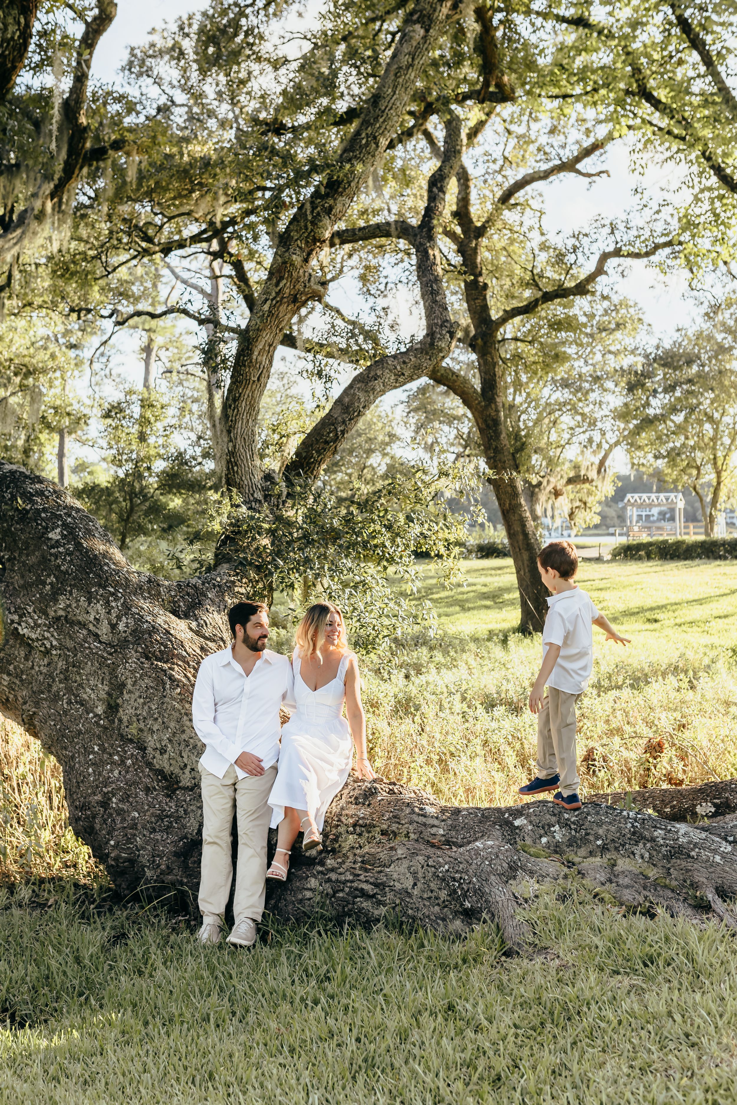 Family of three posing near a large oak tree in a sunlit park, parents in white outfits with young boy standing nearby.