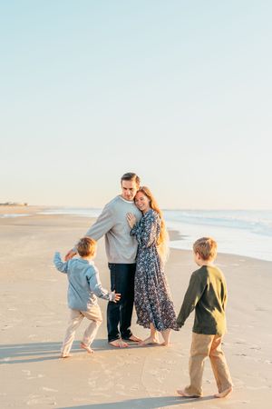 A family in casual clothing walks along a sandy beach at sunset with ocean waves in the background.