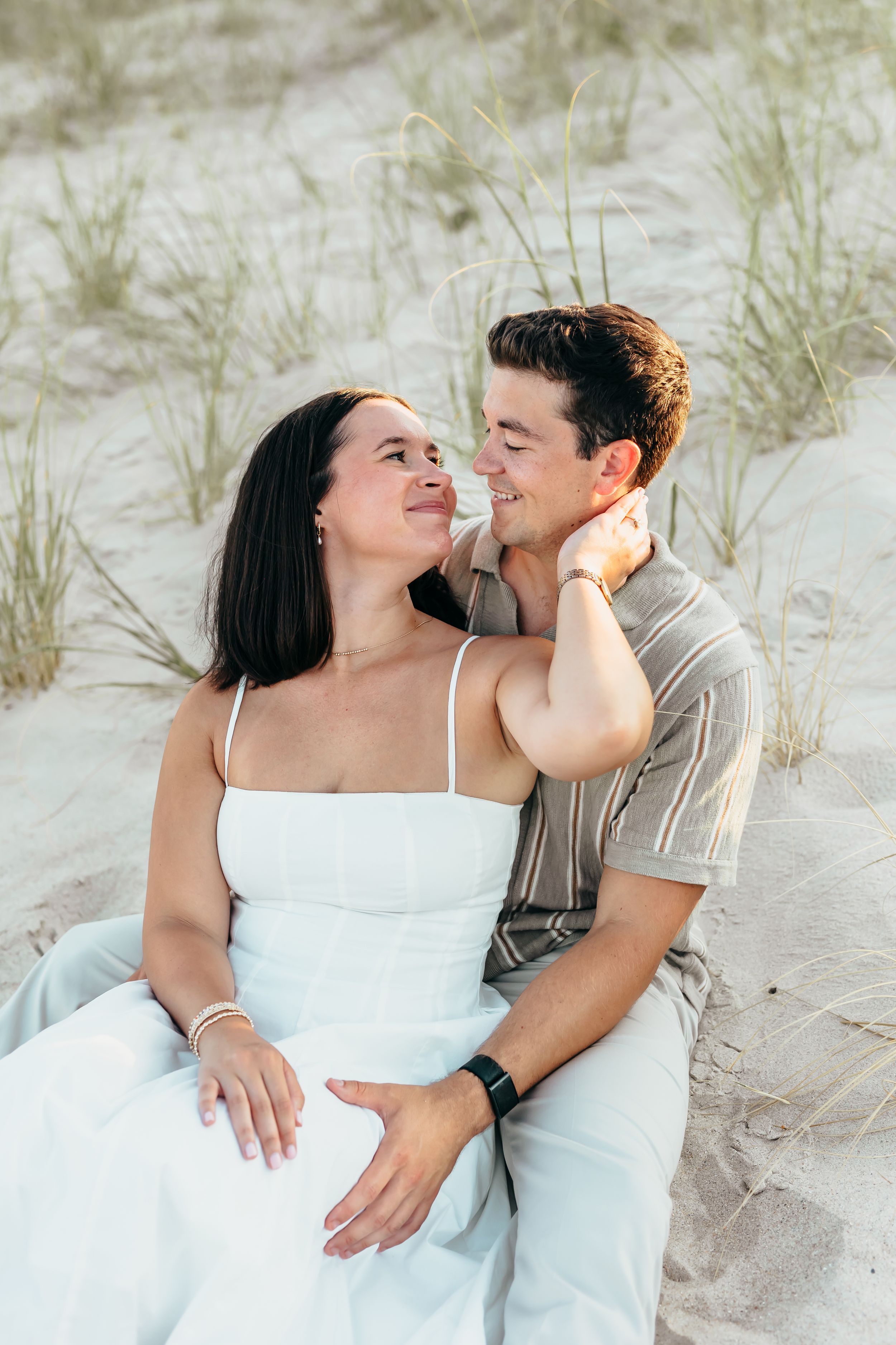 A couple sits together on sandy beach dunes, smiling at each other, woman in white dress, man in striped shirt.