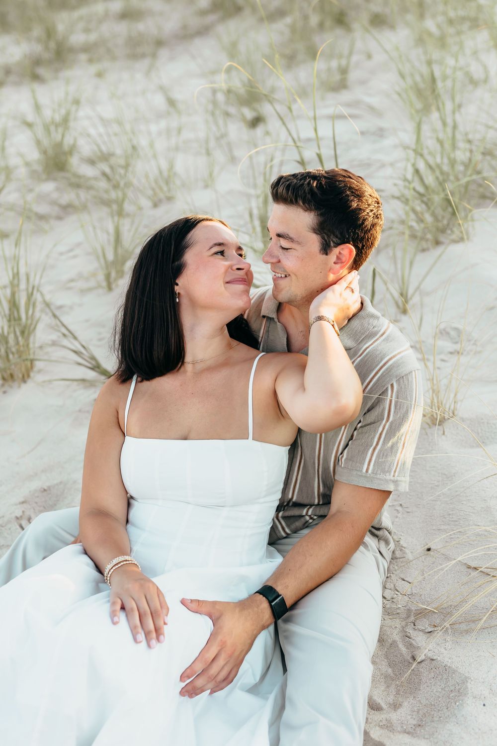 A couple sits together on sandy beach dunes, smiling at each other, woman in white dress, man in striped shirt.