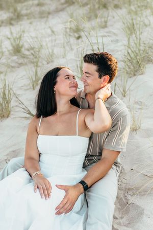A couple sits together on sandy beach dunes, smiling at each other, woman in white dress, man in striped shirt.