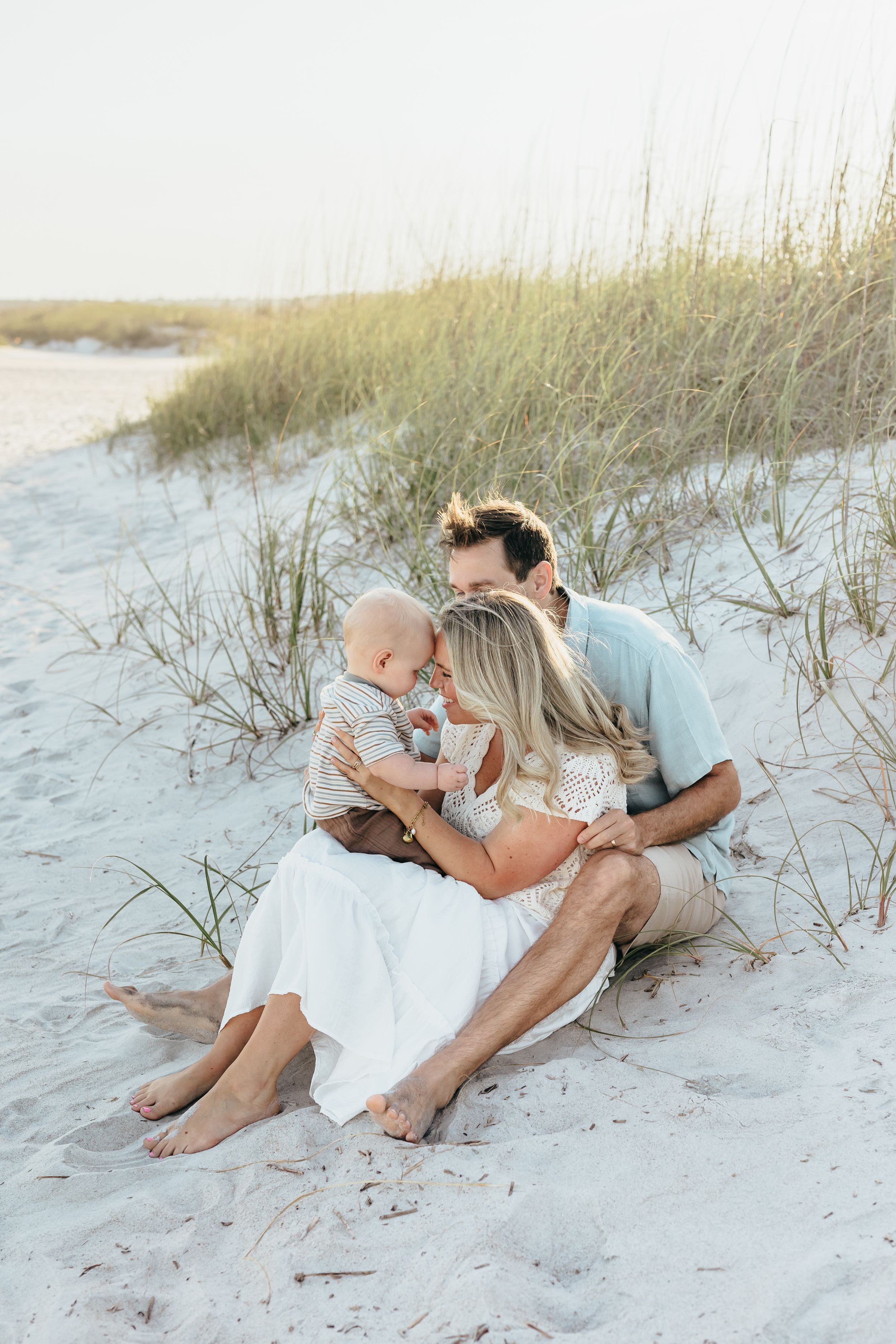 A young family sits together on white sand dunes, parents lovingly nuzzling their baby boy near beach grass.