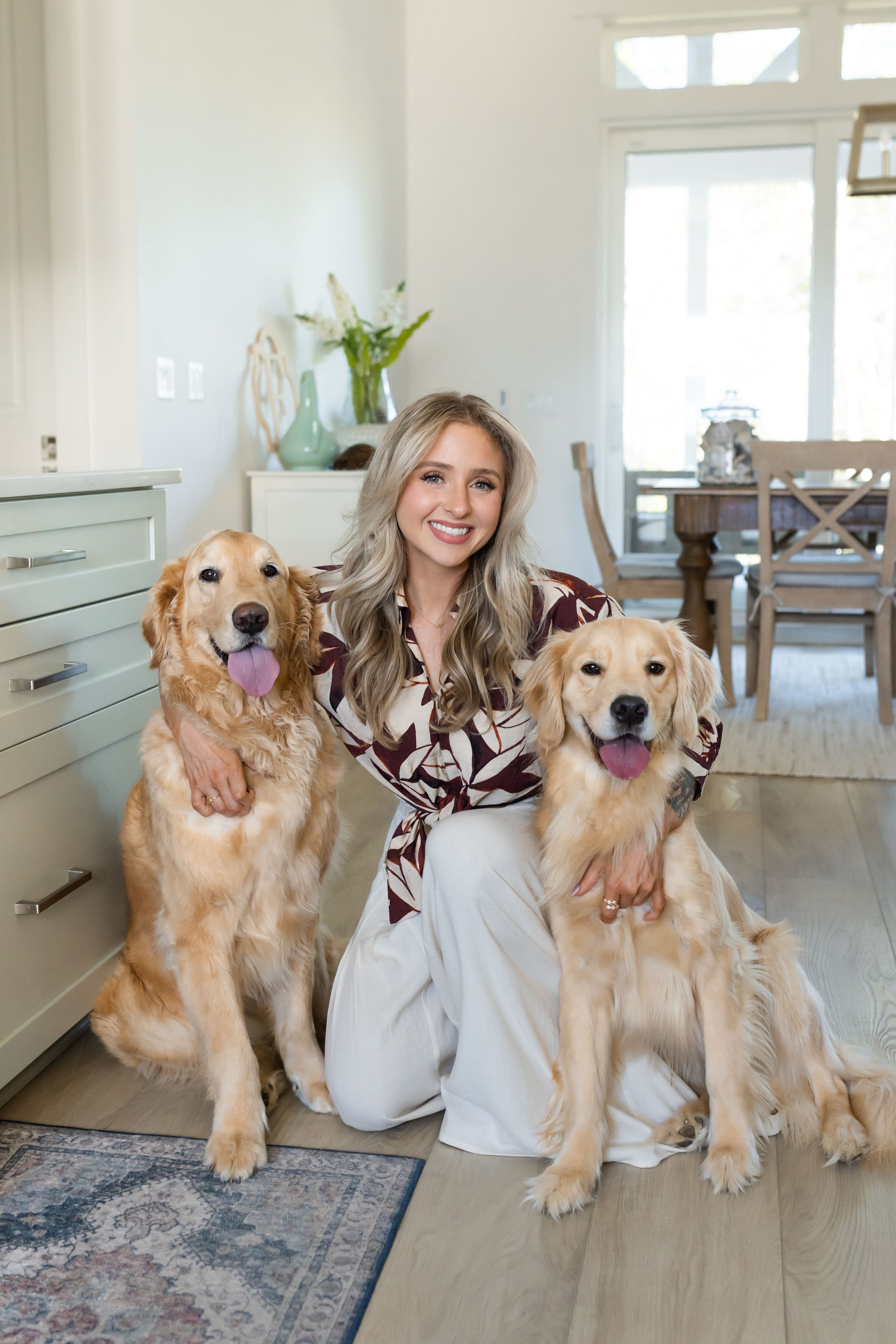 Smiling blonde woman posing with two golden retrievers on hardwood floor in bright modern home interior.