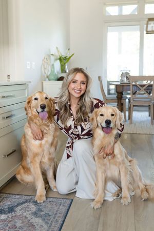 Smiling blonde woman posing with two golden retrievers on hardwood floor in bright modern home interior.