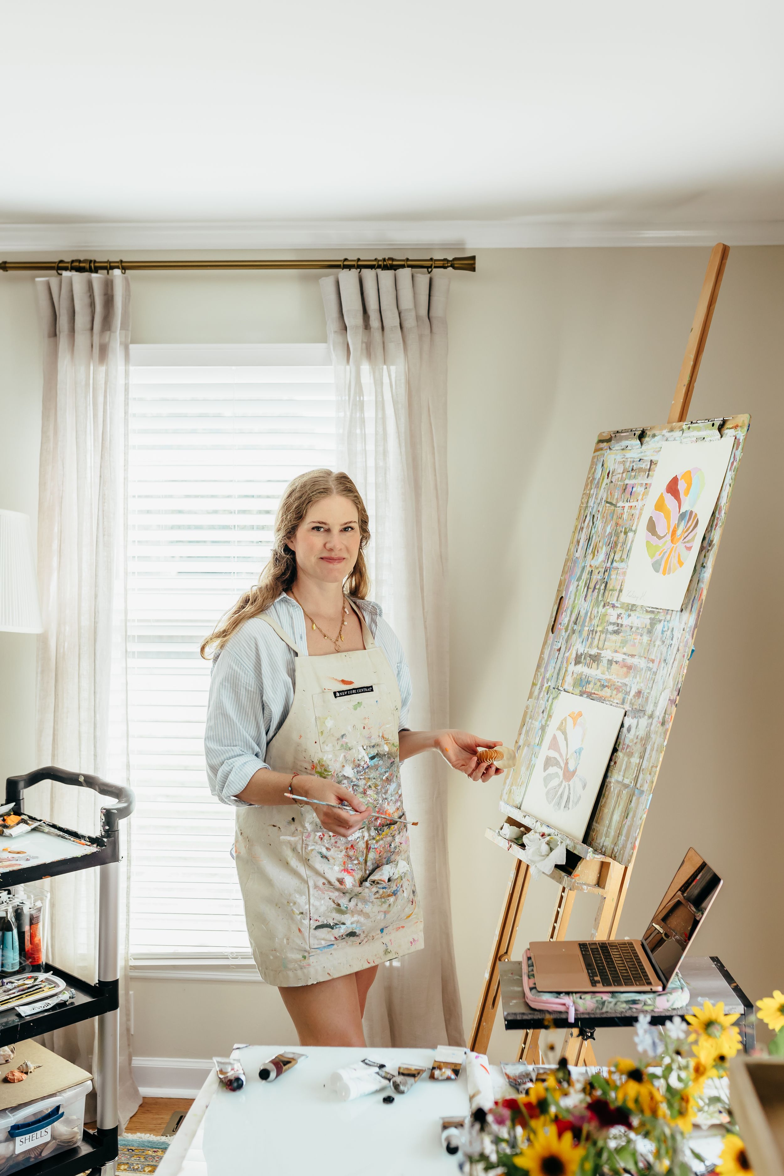 Artist in paint-splattered apron stands by wooden easel with colorful artwork in bright home studio.