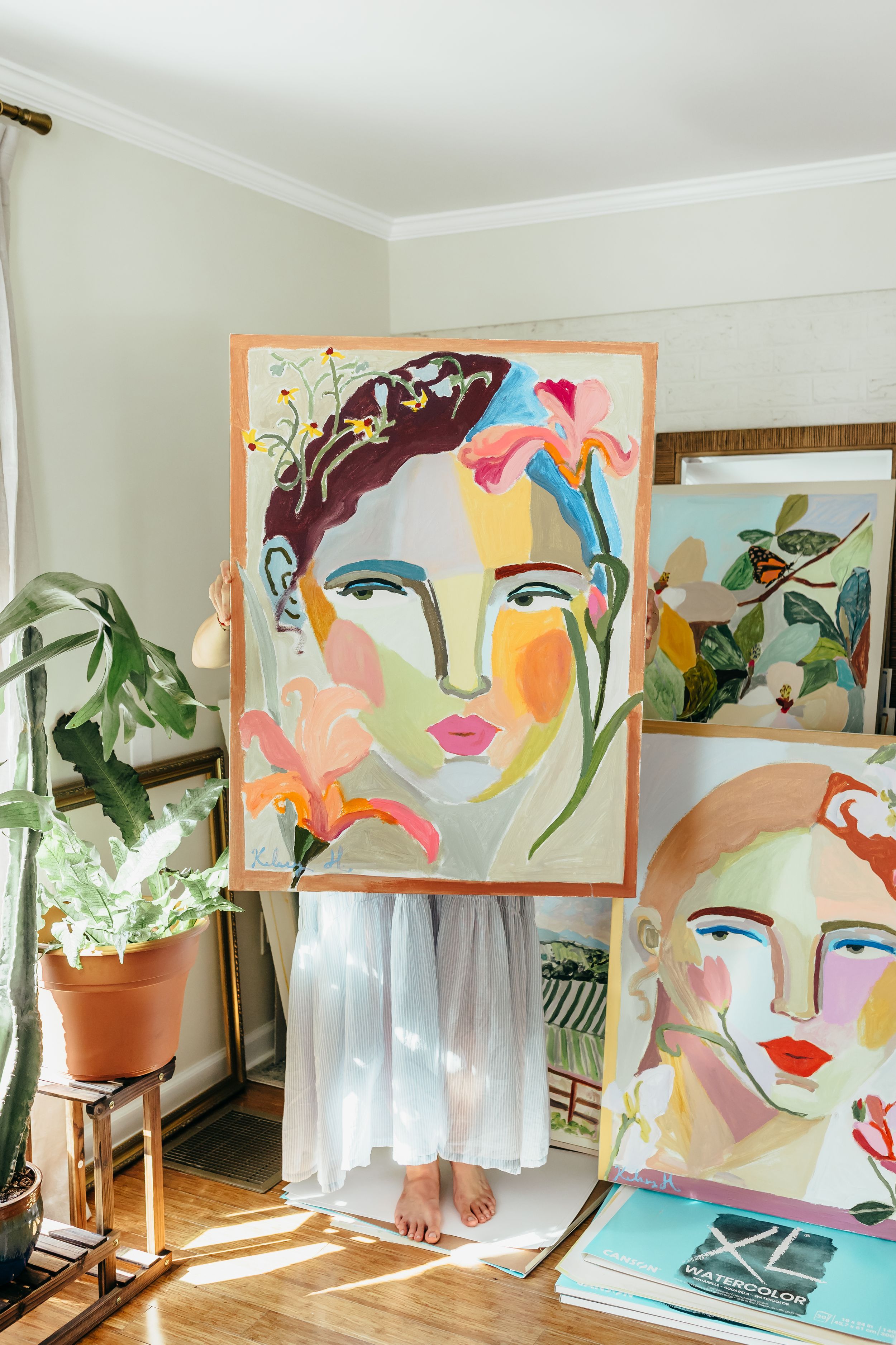 Artist holding a colorful portrait painting in a bright studio with plants and artwork displayed nearby.