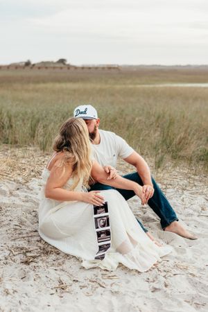 Couple sitting on beach sand with ultrasound photos, sharing an intimate moment at sunset.