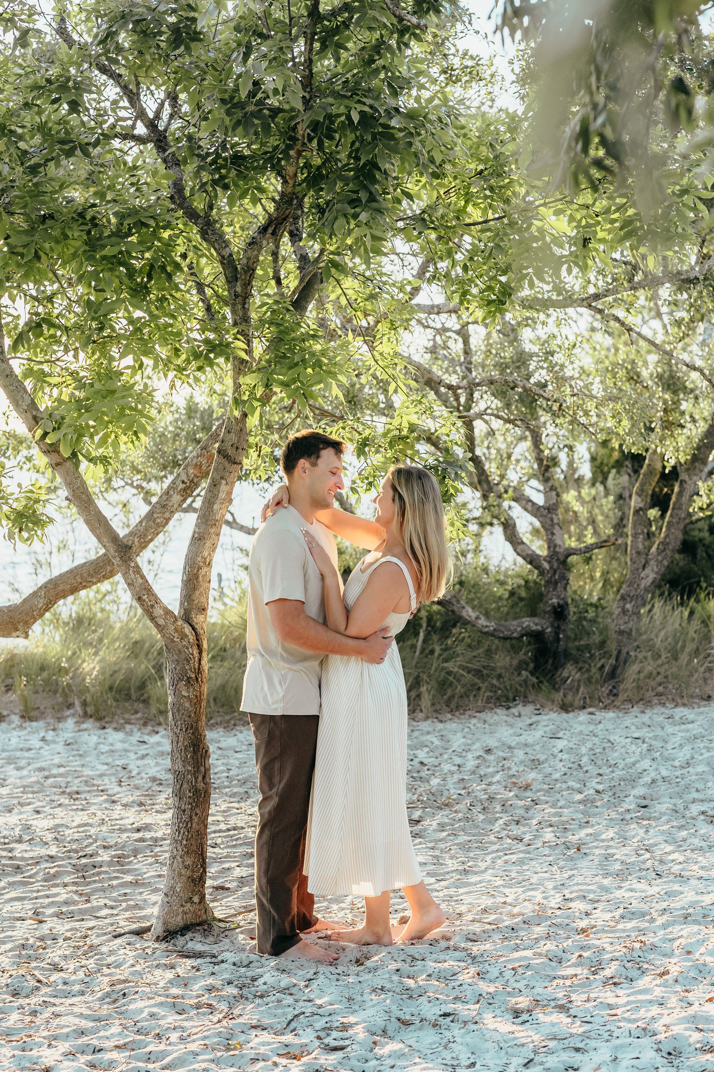 Couple embracing under green trees on sandy beach, woman in white dress and man in casual attire, golden sunlight filtering through.